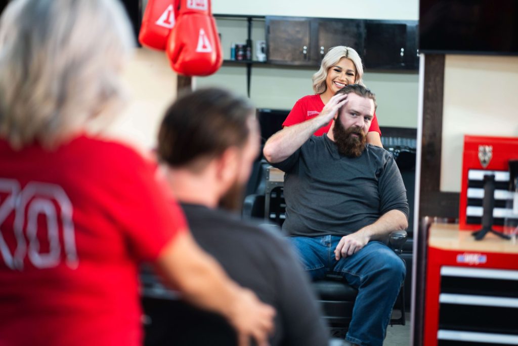 Knockouts Arlington client with fresh cut and trimmed beard sits in barber chair with a Knockouts stylist standing behind him. Both face a mirror appreciating the finished haircut.